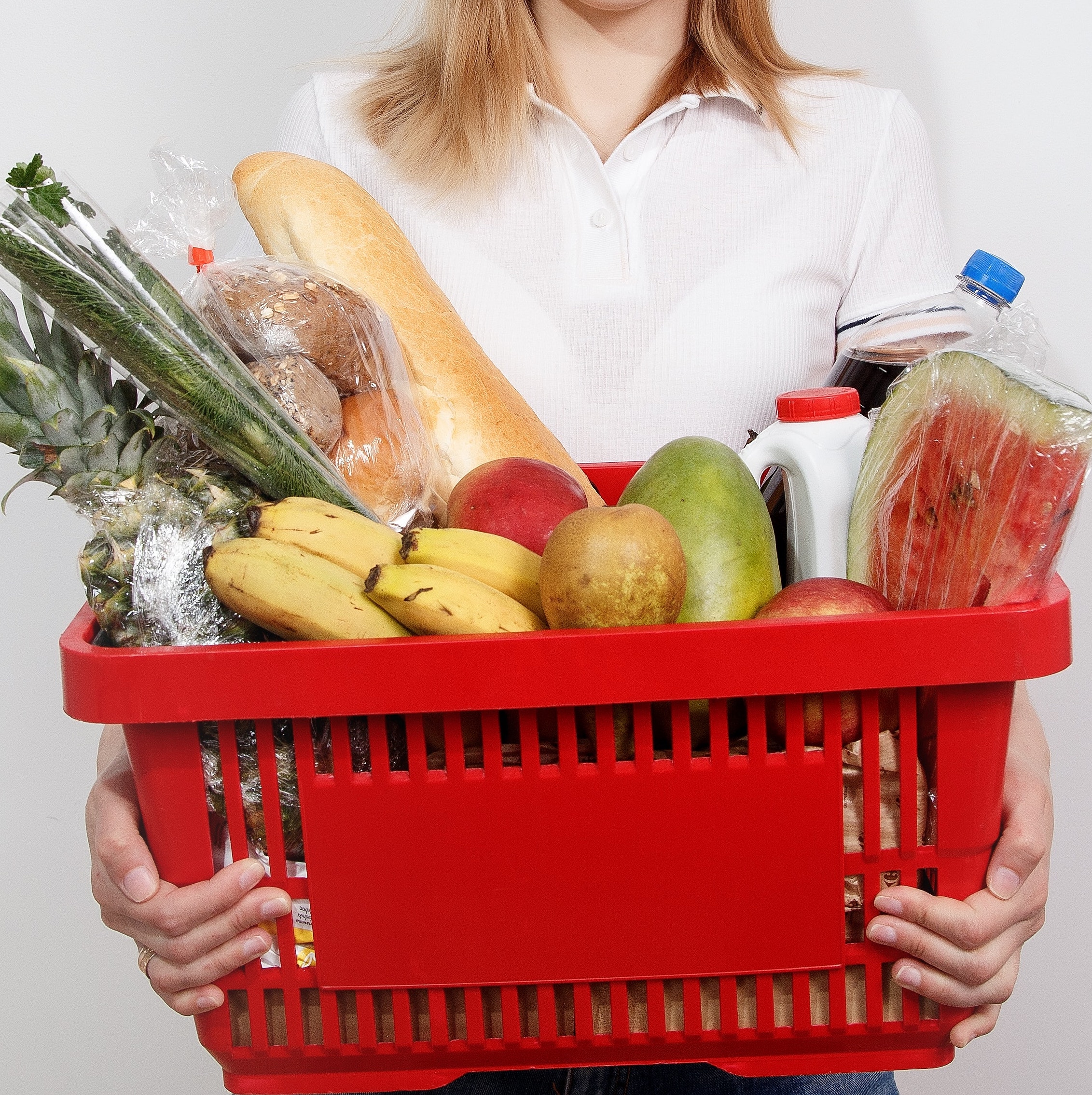 A red shopping basket full of bread, fruit and bottles, being held by a person wearing a white shirt.