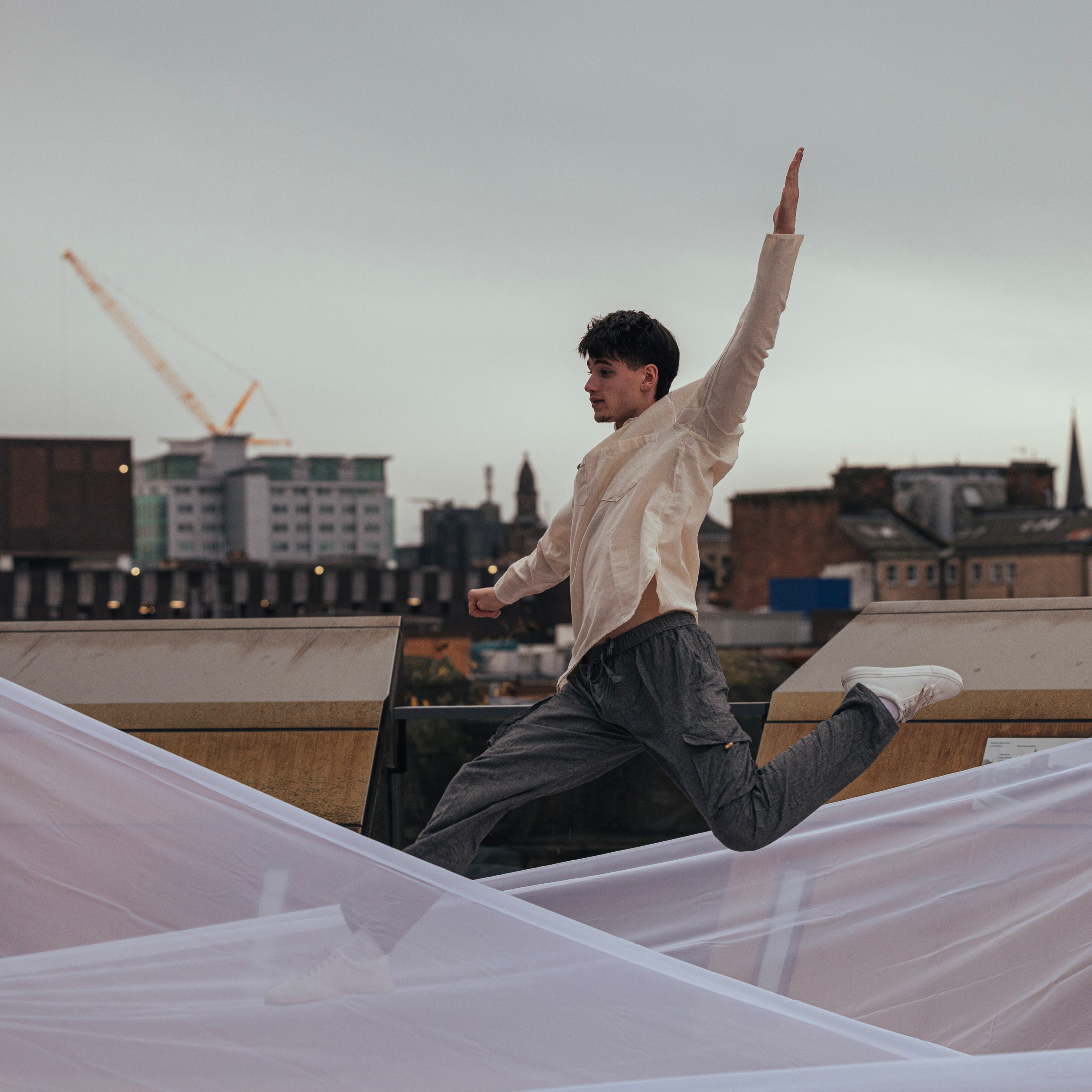 Photo of a young man dancing. He is mid-leap with one arm in the air. He is on a roof top with buildings in the background, and there are netted sheets surrounding him.