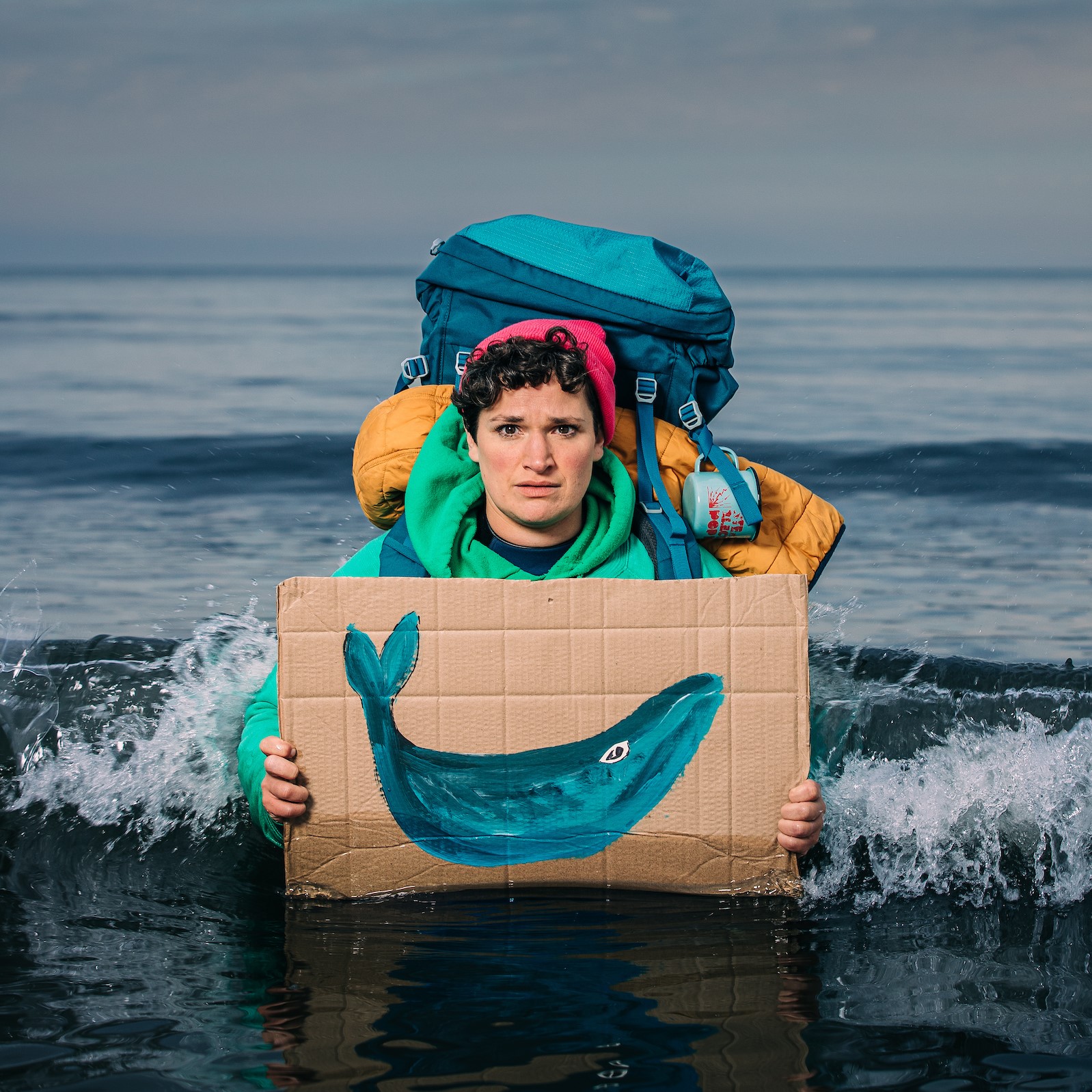 A woman wearing a large rucksack and holding a cardboard sign with a whale painted on it. She is standing up to her waist in the sea and has a concerned expression
