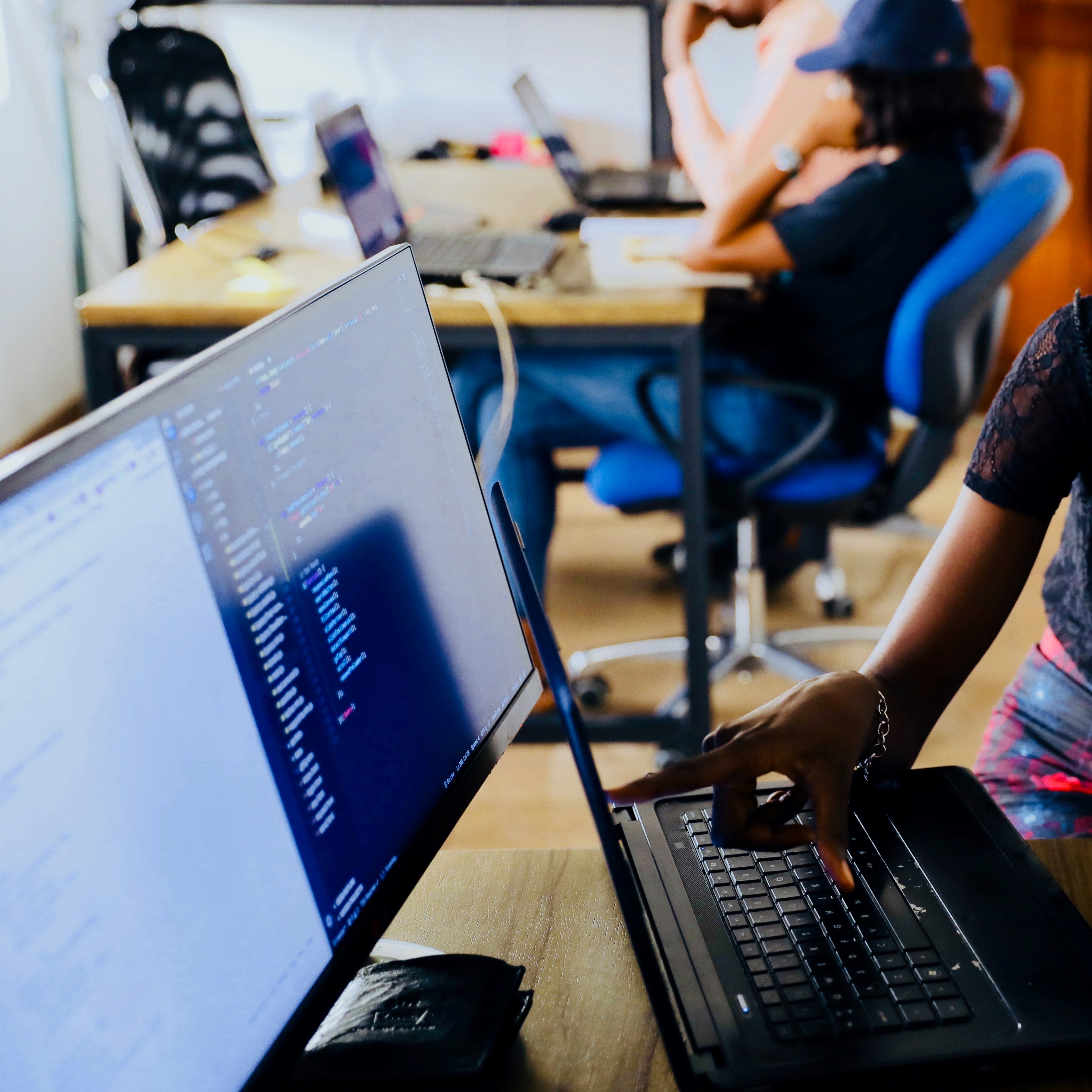 A computer screen with a coding programme open. In front of that is a laptop, with a woman's hand and arm in shot, pointing at something on its screen