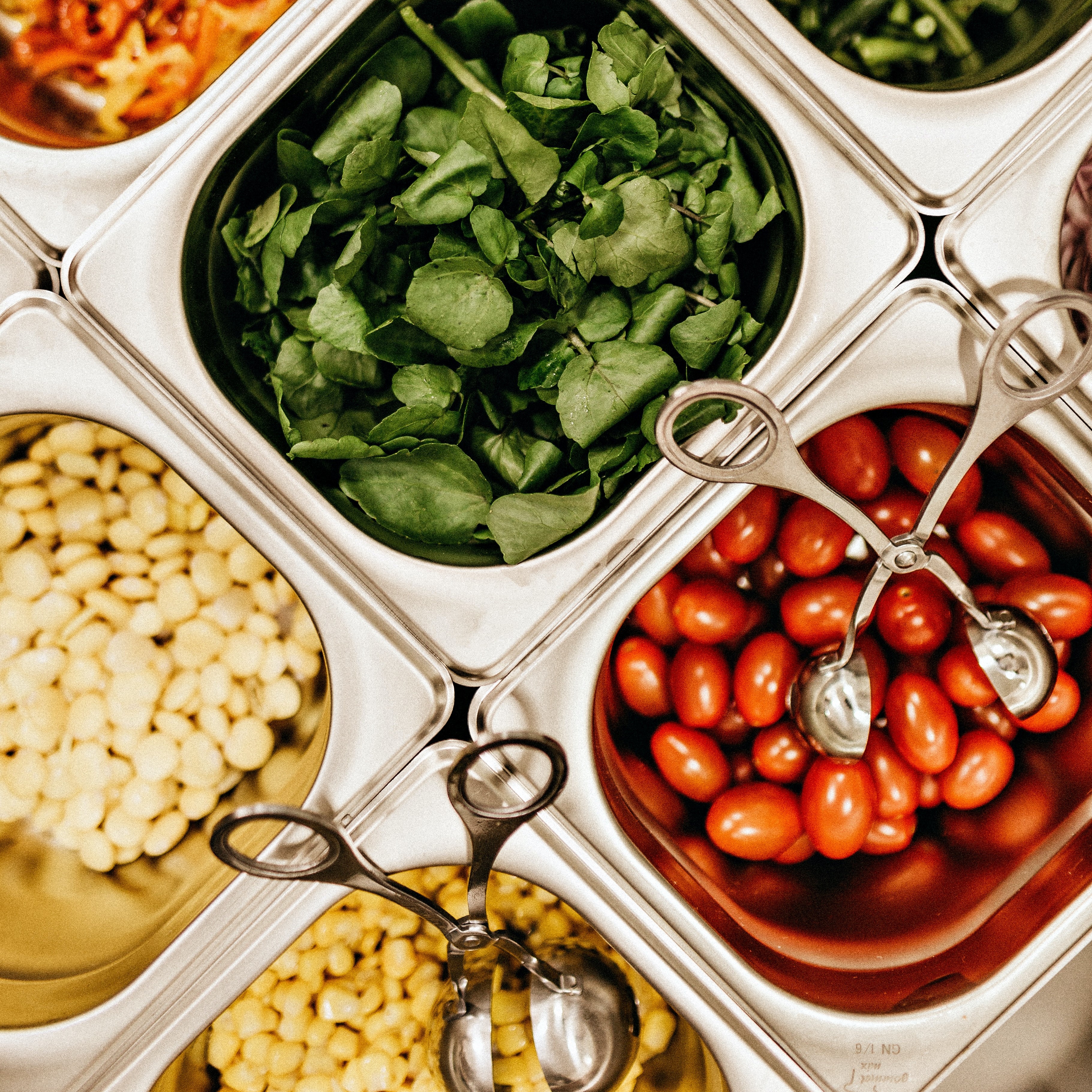 Photo of metal food containers holding salad leaves, sweetcorn and tomatoes, with utensils in the dishes.