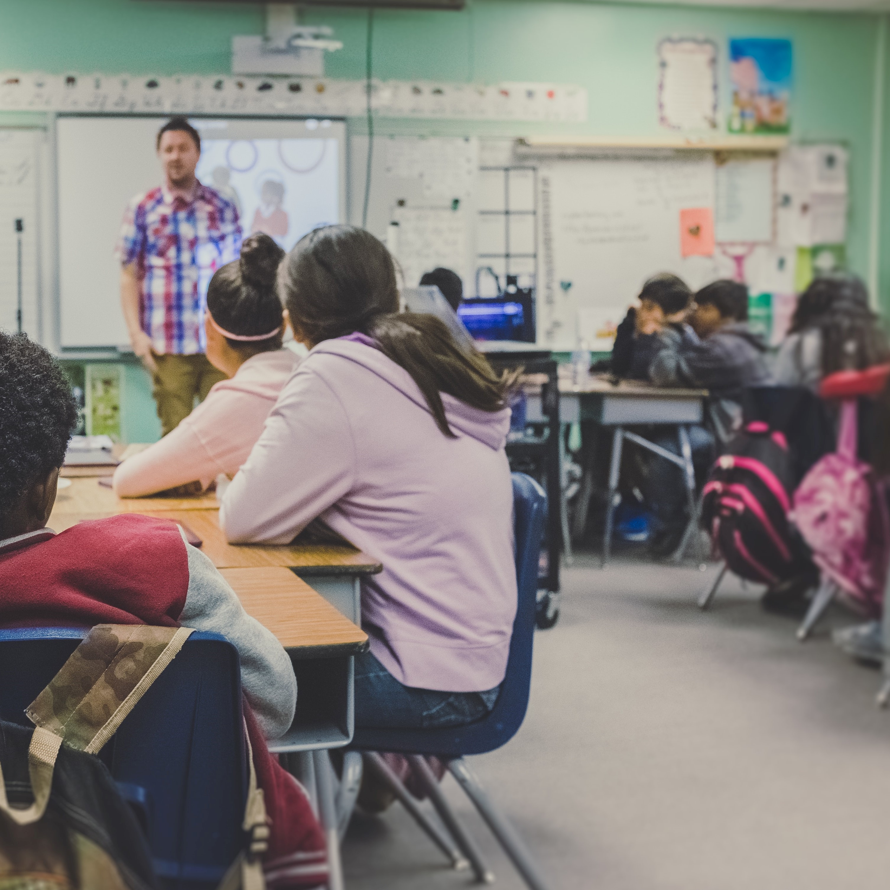 Photo of school classroom, with children at tables looking towards a teacher who is speaking at the front