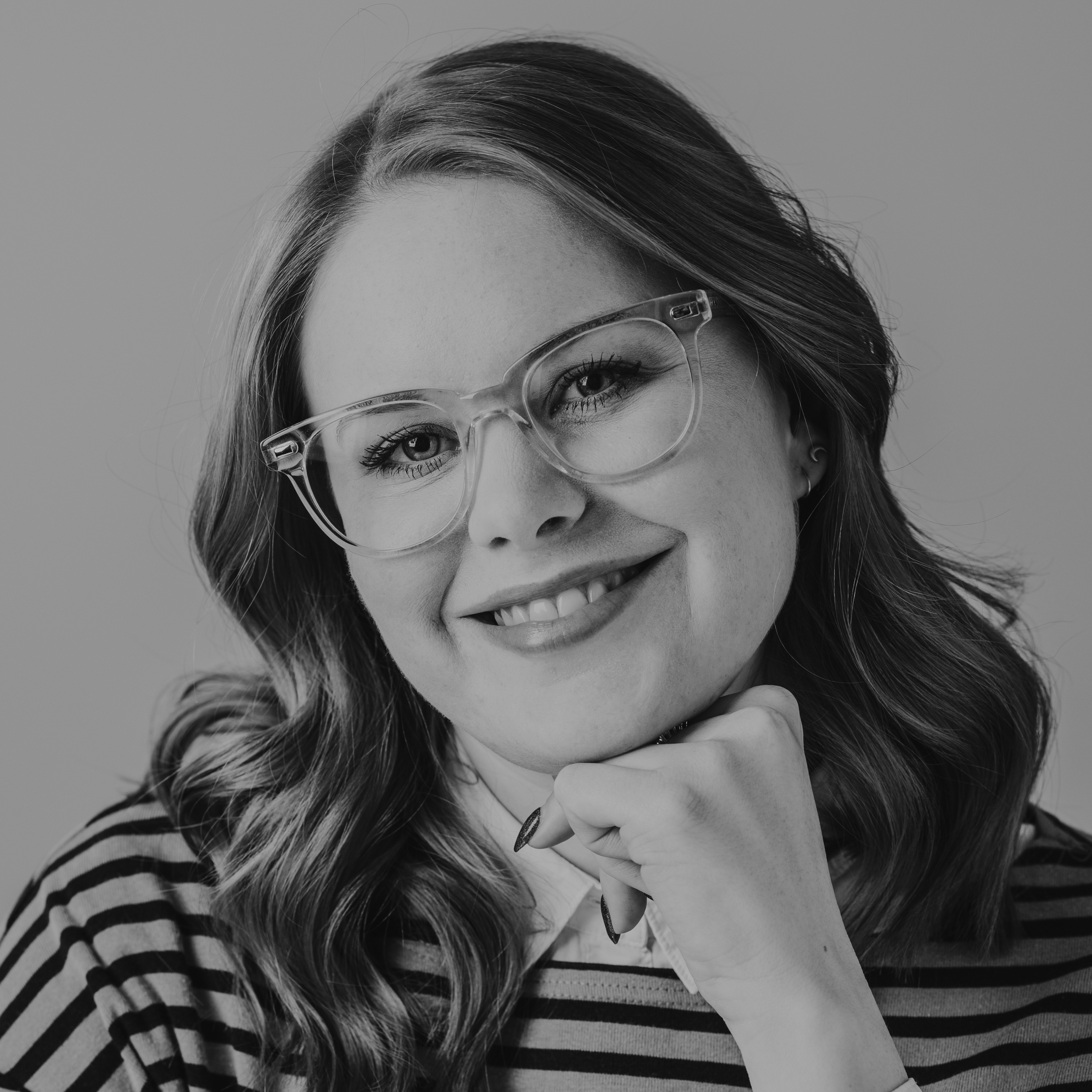 A greyscale image of a person wearing glasses. They have long hair parted in the centre and wear glasses, Their chin rests on their hand and they are wearing a striped top