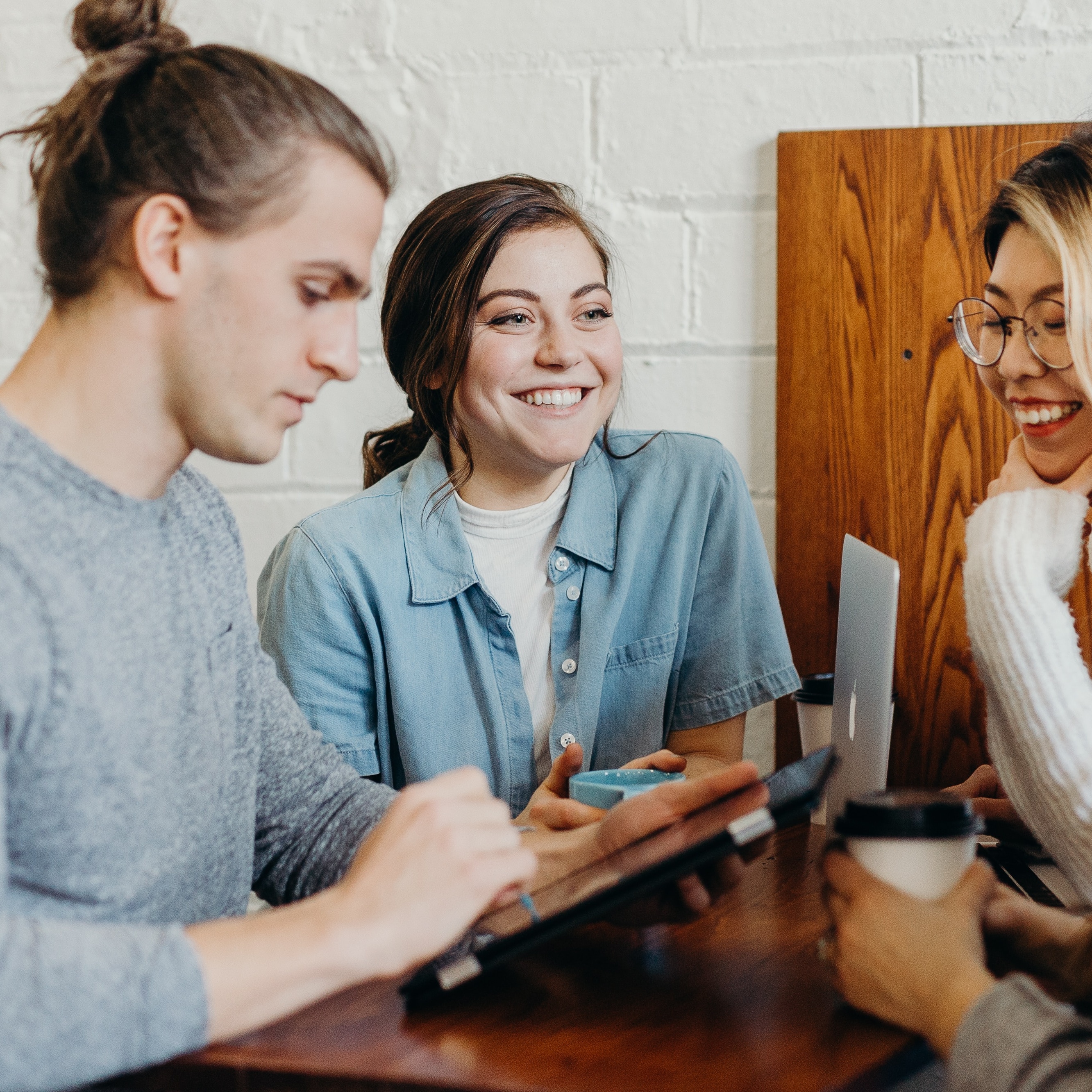 Three young adults stand around a table. Two are smiling one is looking down at a tablet.