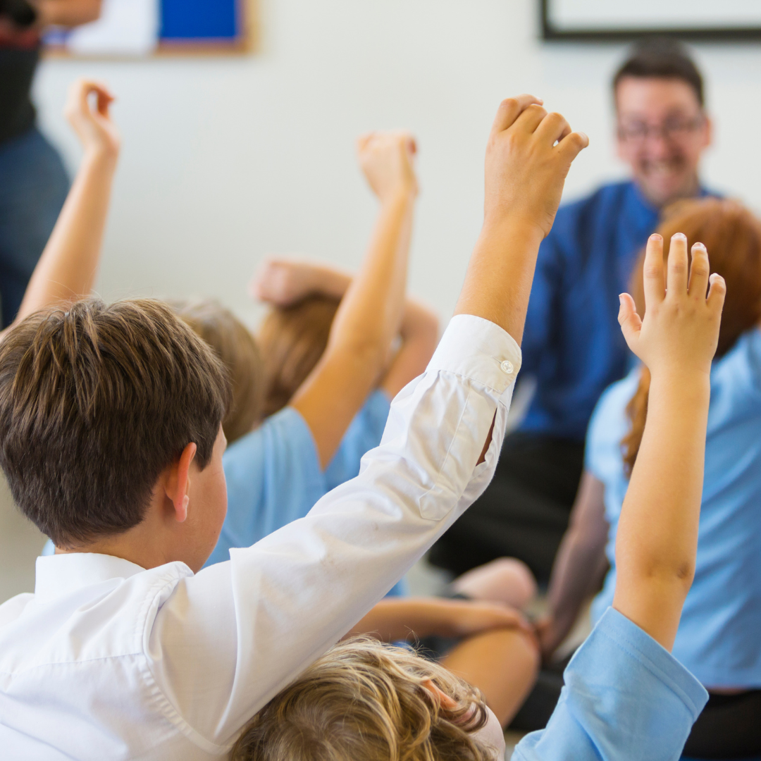 School children raise their hands in the classroom with teacher in the background