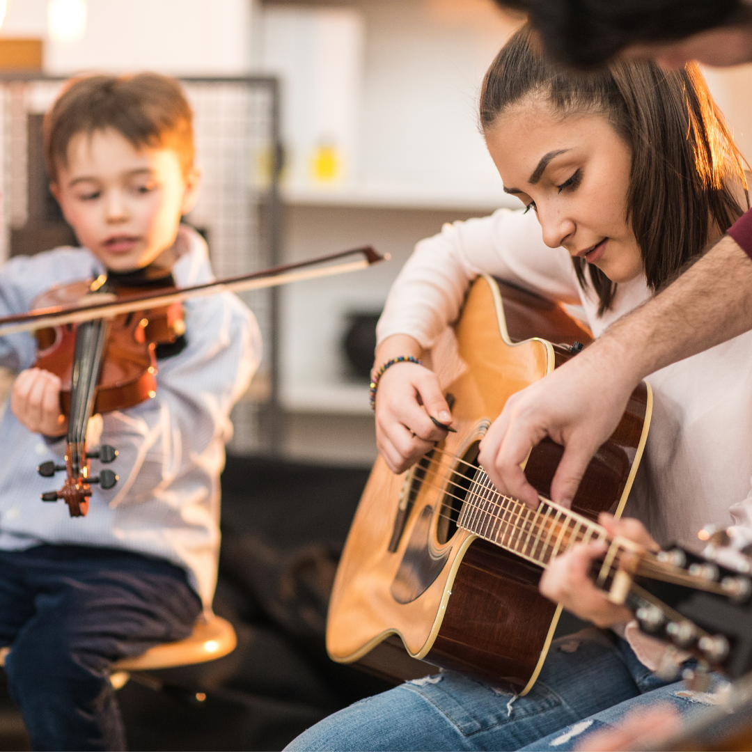 A young person plays guitar in the foreground, while another plays violin in the background