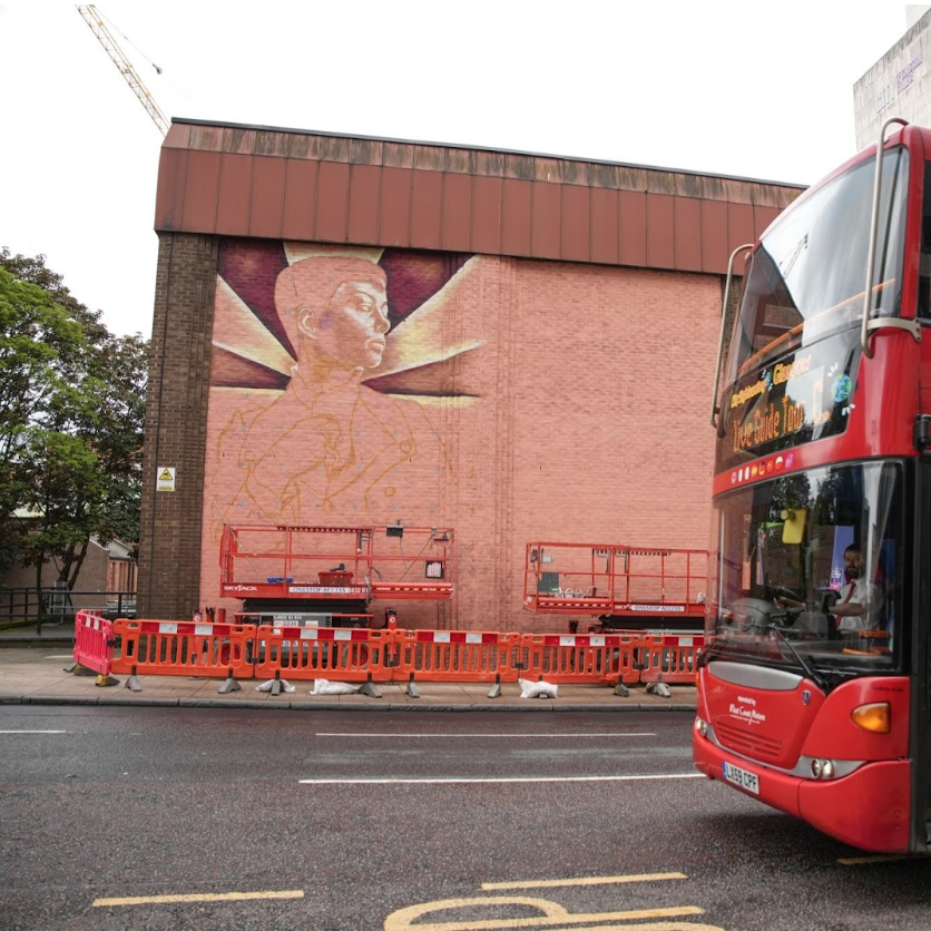 A street mural take shape on a building wall, a bus driving past in the foreground