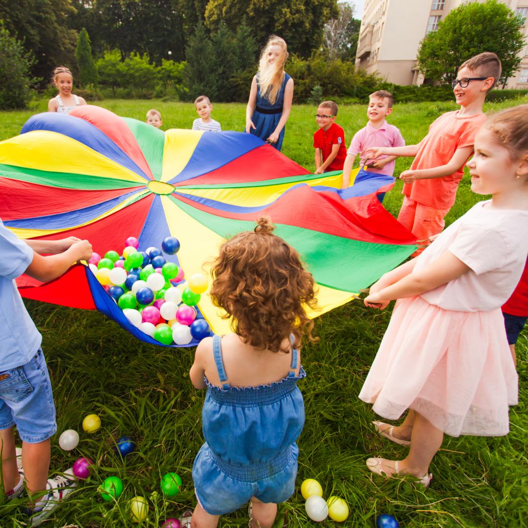 A group of young children play outside on grass with a multi-coloured parachute