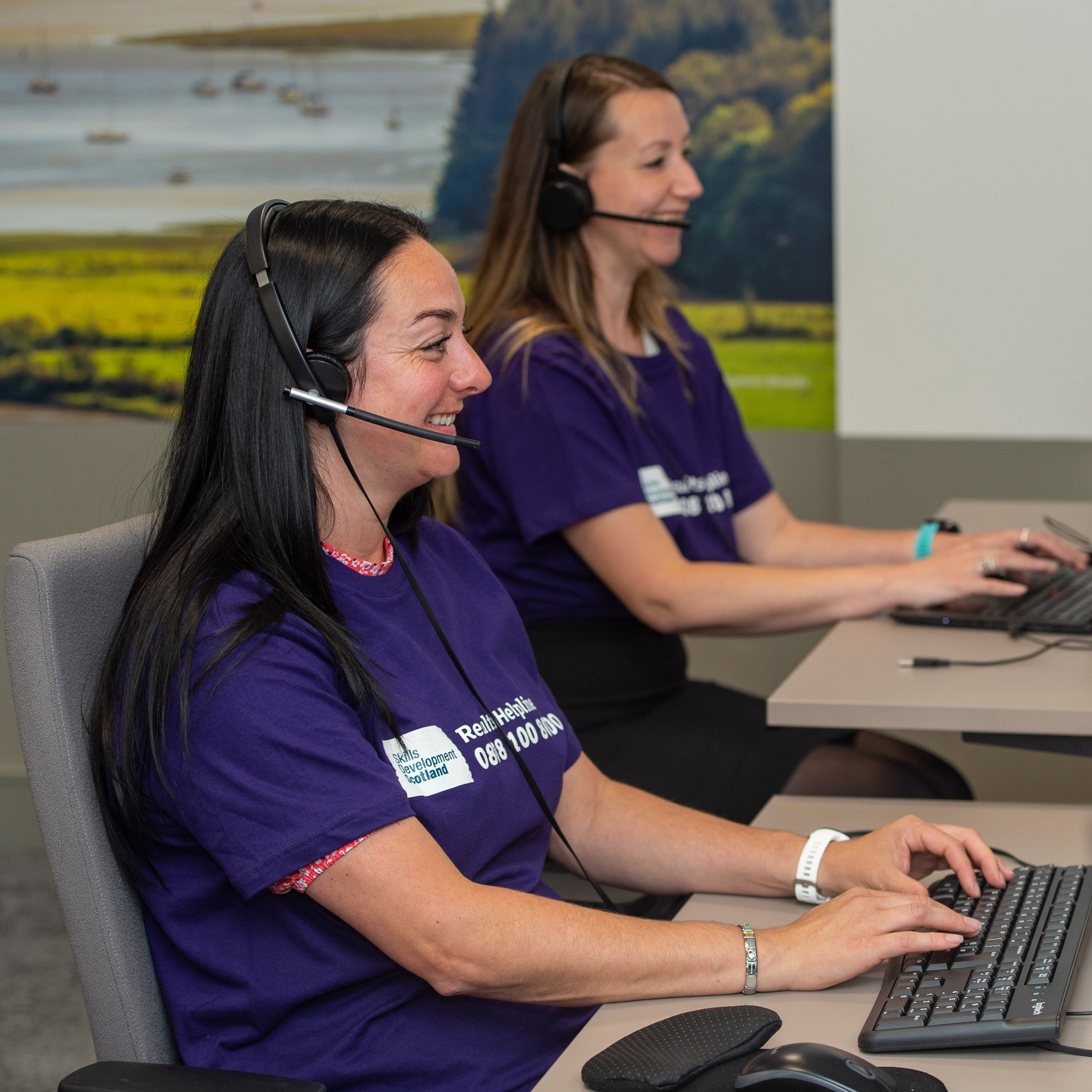 Two smiling people wear purple t-shirts and headsets while typing at keyboards