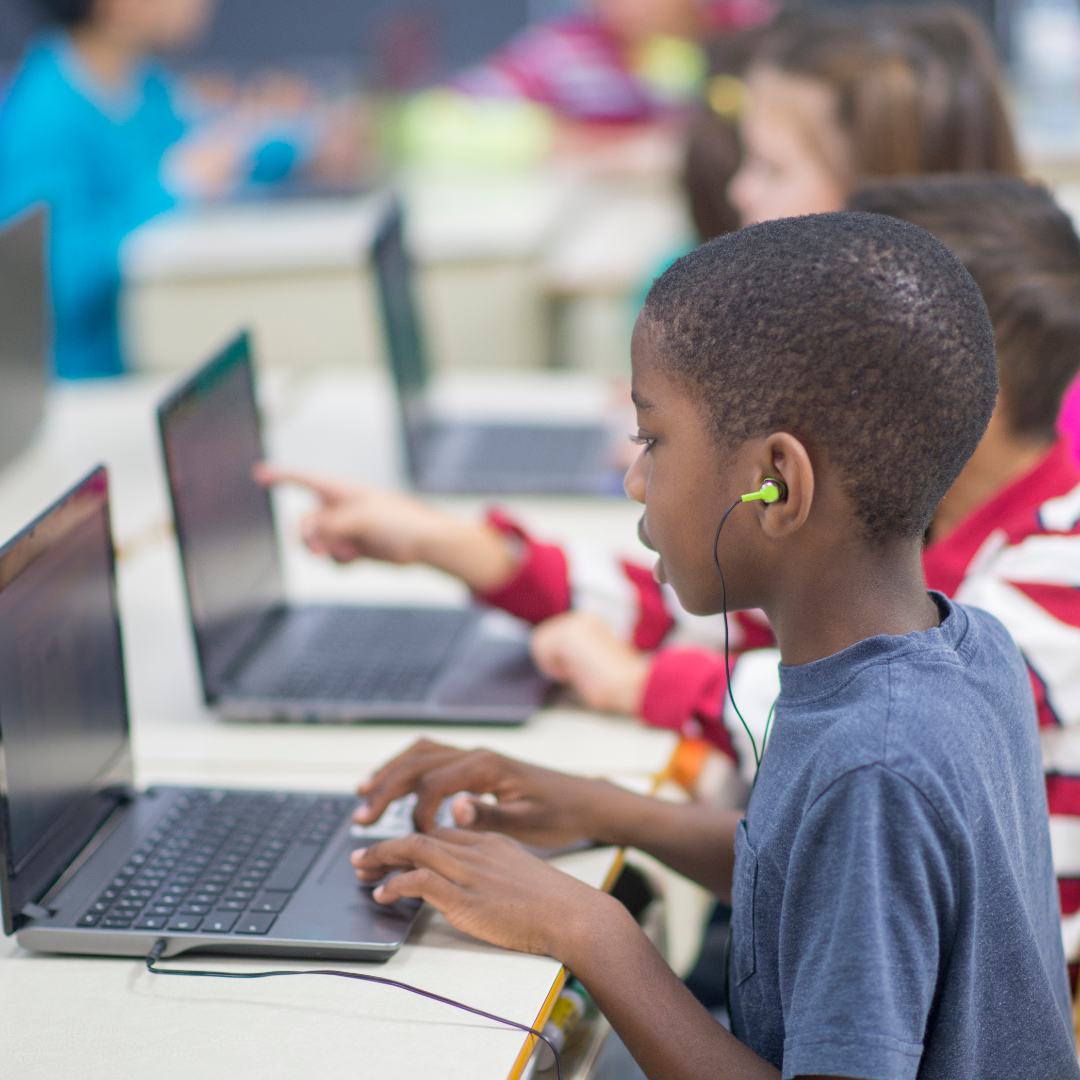 A child sits at a laptop. They have bright green earbuds in. More children seated at laptops are visible in the background