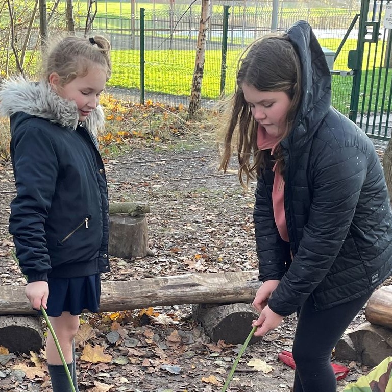 Two children in outdoor coats each holding a stick and playing in leaves