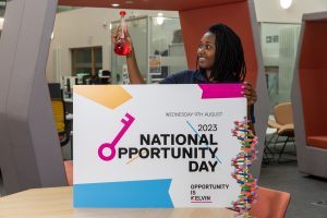 A young woman holds up a glass beaker filled with red liquid. In her other hand, she holds a sign that reads National Opportunity Day.