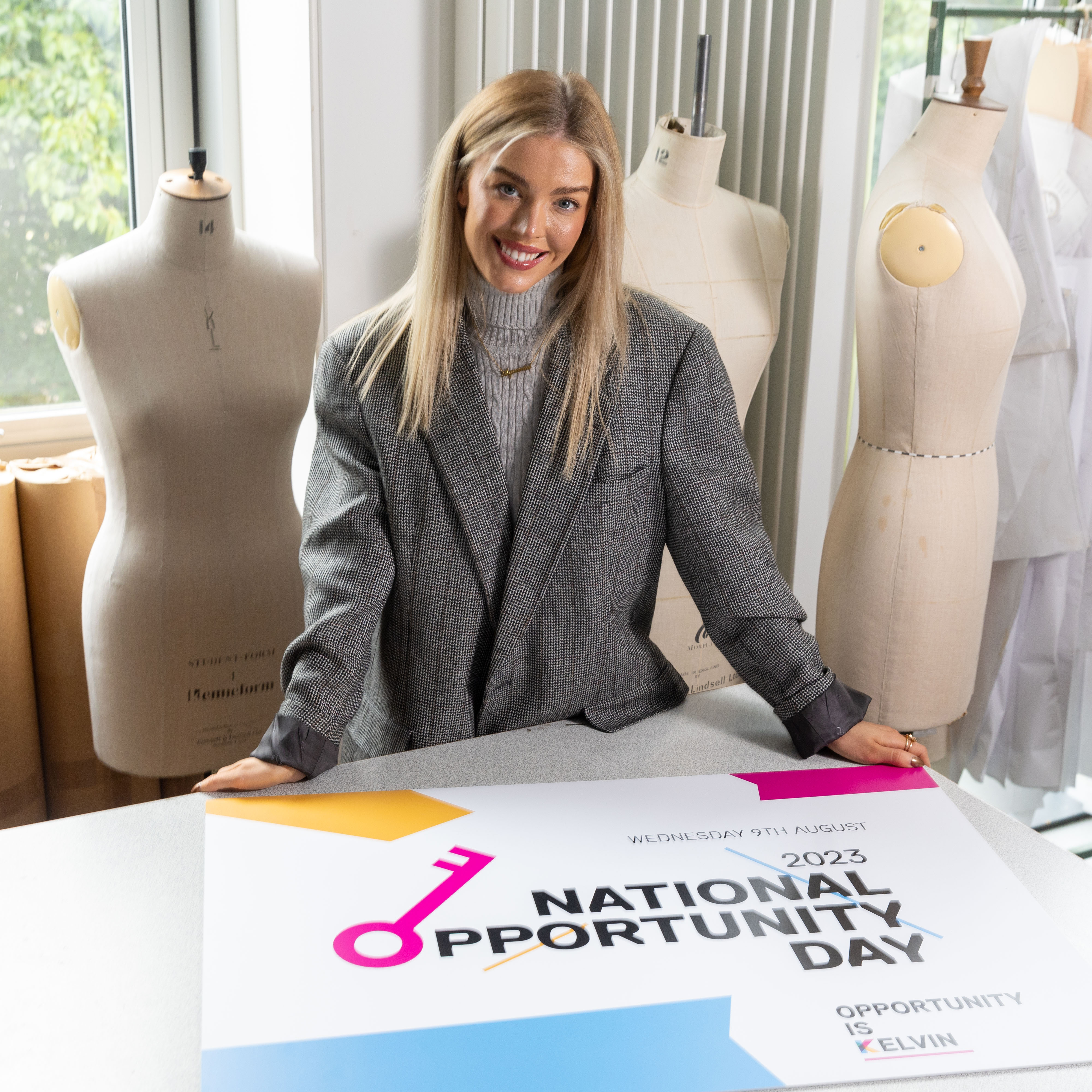 A person with long blonde hair stands in front of dressmaking mannequins. In front of her on a table, a sign reads National Opportunity Day