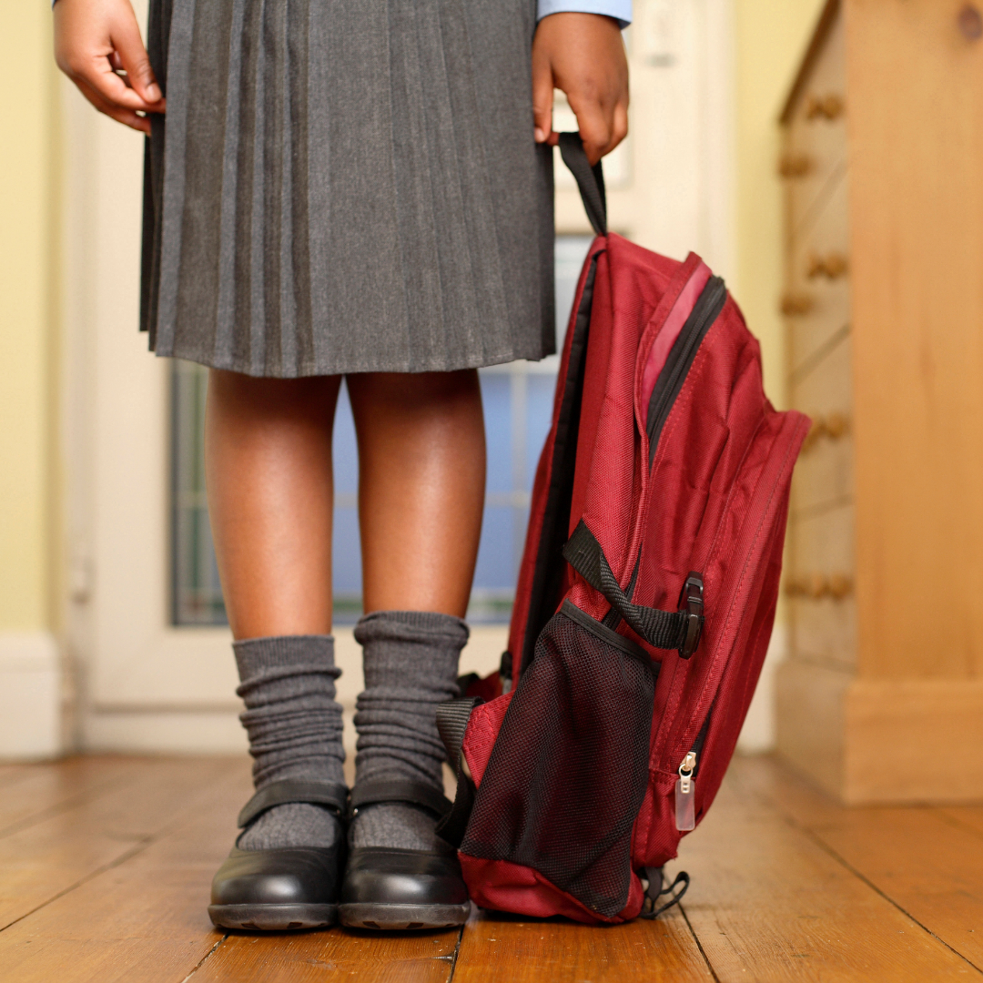 A small child dressed in school uniform is pictured holding a red backpack. They are wearing a grey pleated skirt, grey socks and black shoes, with only their legs and hands shown in the frame.