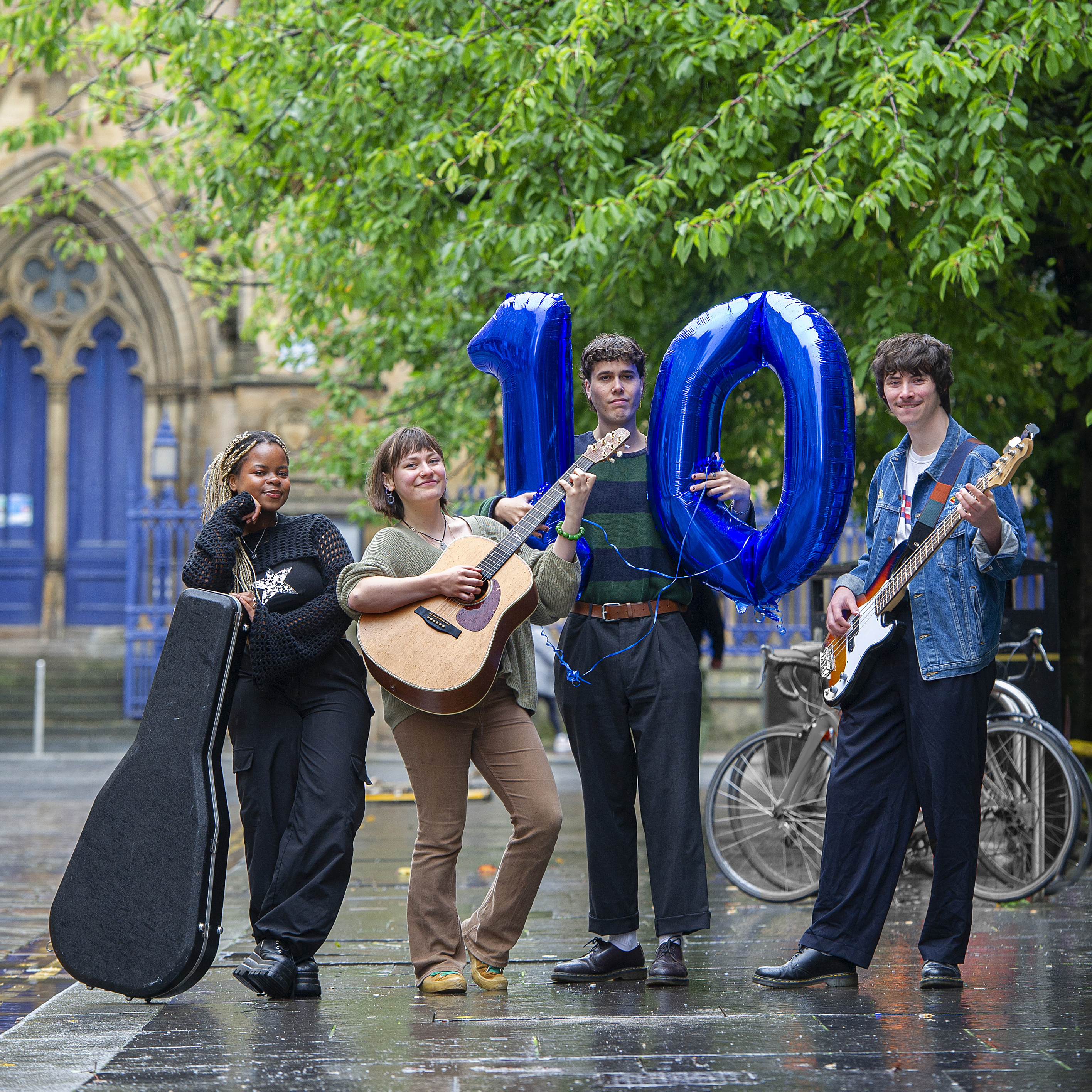 Four people stand outside in front of leafy green trees. They hold guitars, while another has two blue balloons in the shape of a one and zero