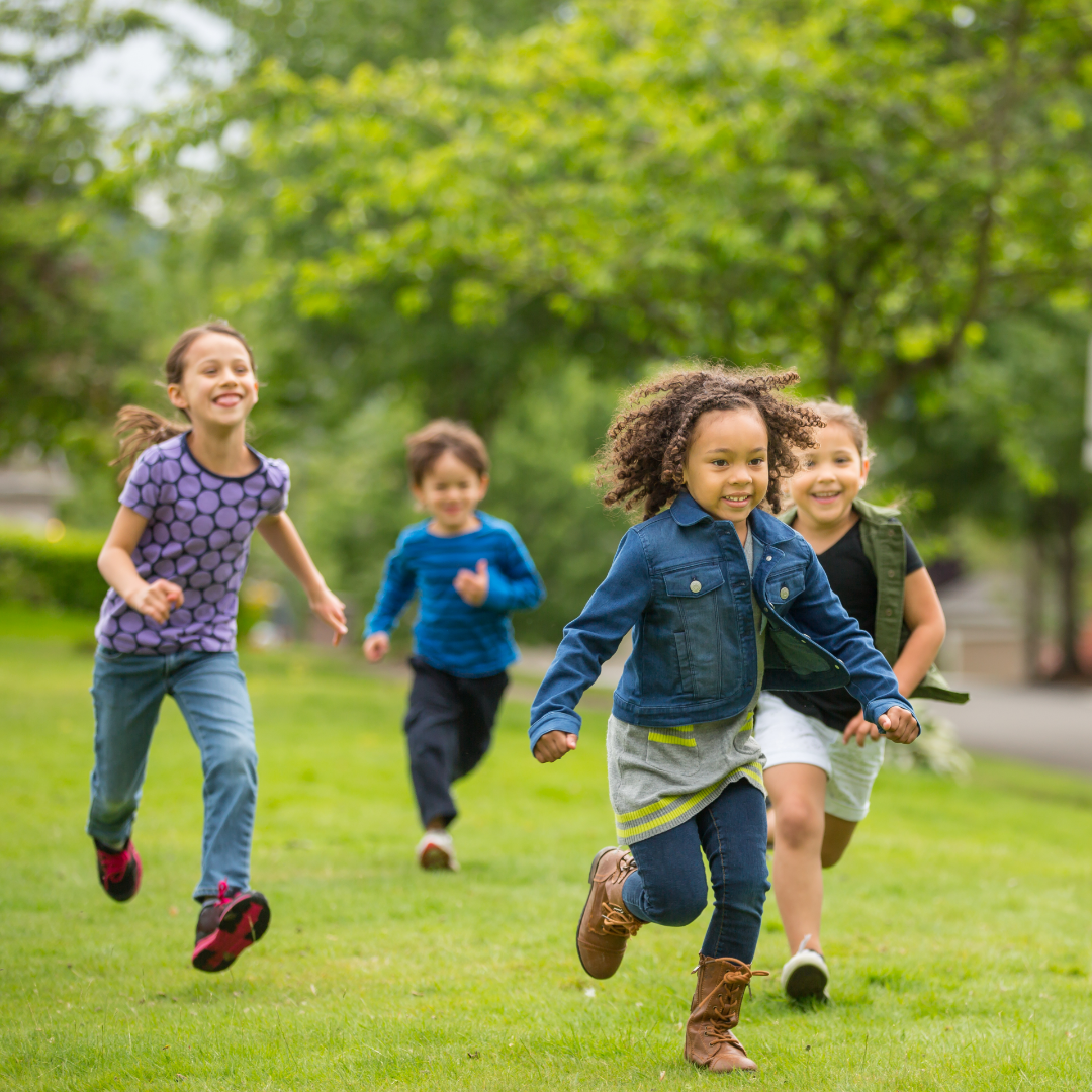 Four young children running outdoors. There is grass under their feet and trees are visible in the background