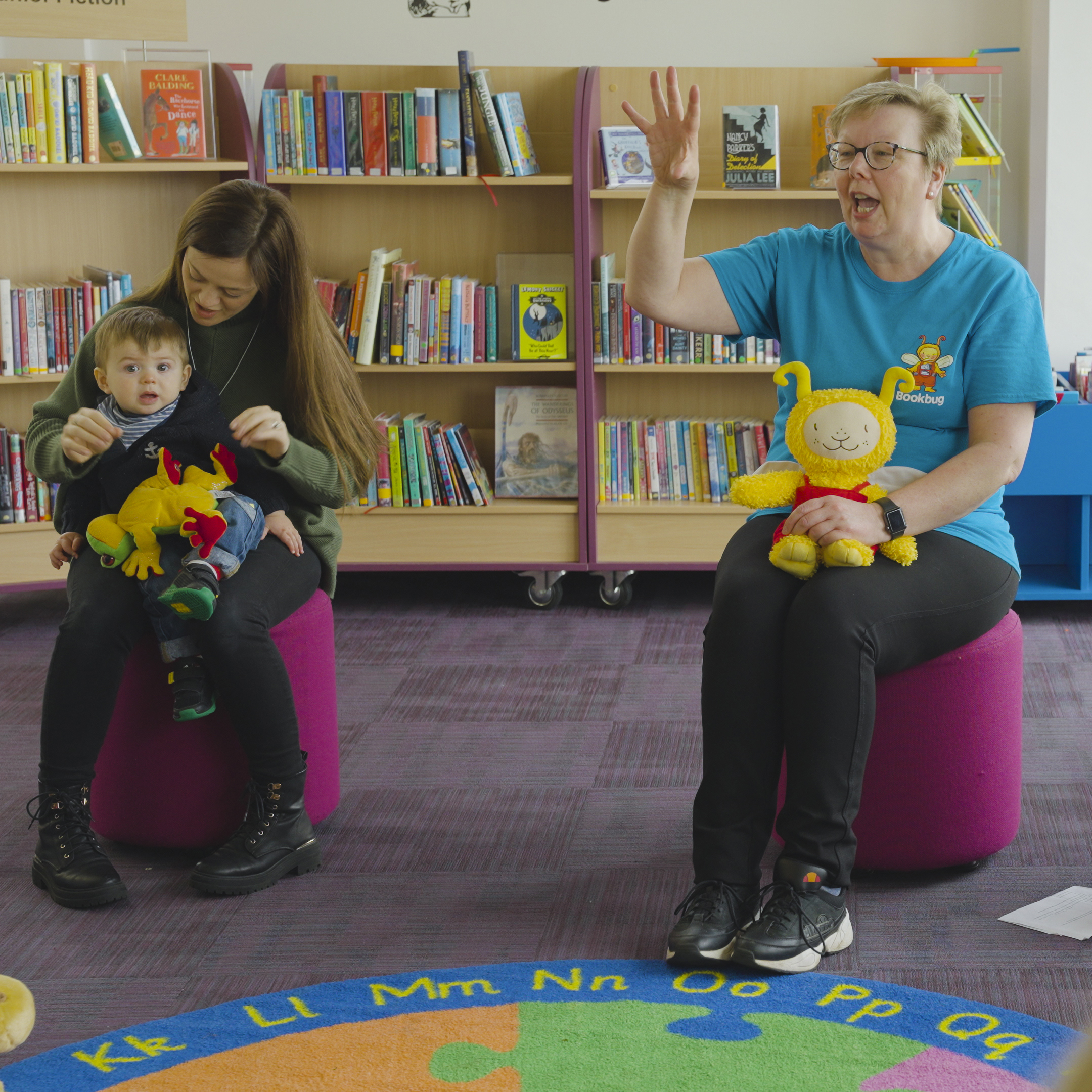 Two people sitting on purple stools in front of shelves of books. The person on the right has a yellow soft toy sitting on their lap. The person on the left has a small child sitting on their lap.