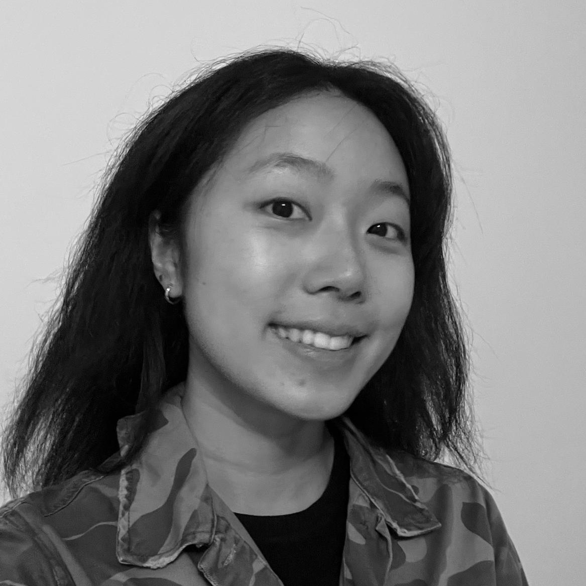 A greyscale image of a smiling person with long dark hair and wearing a camoflage print jacket
