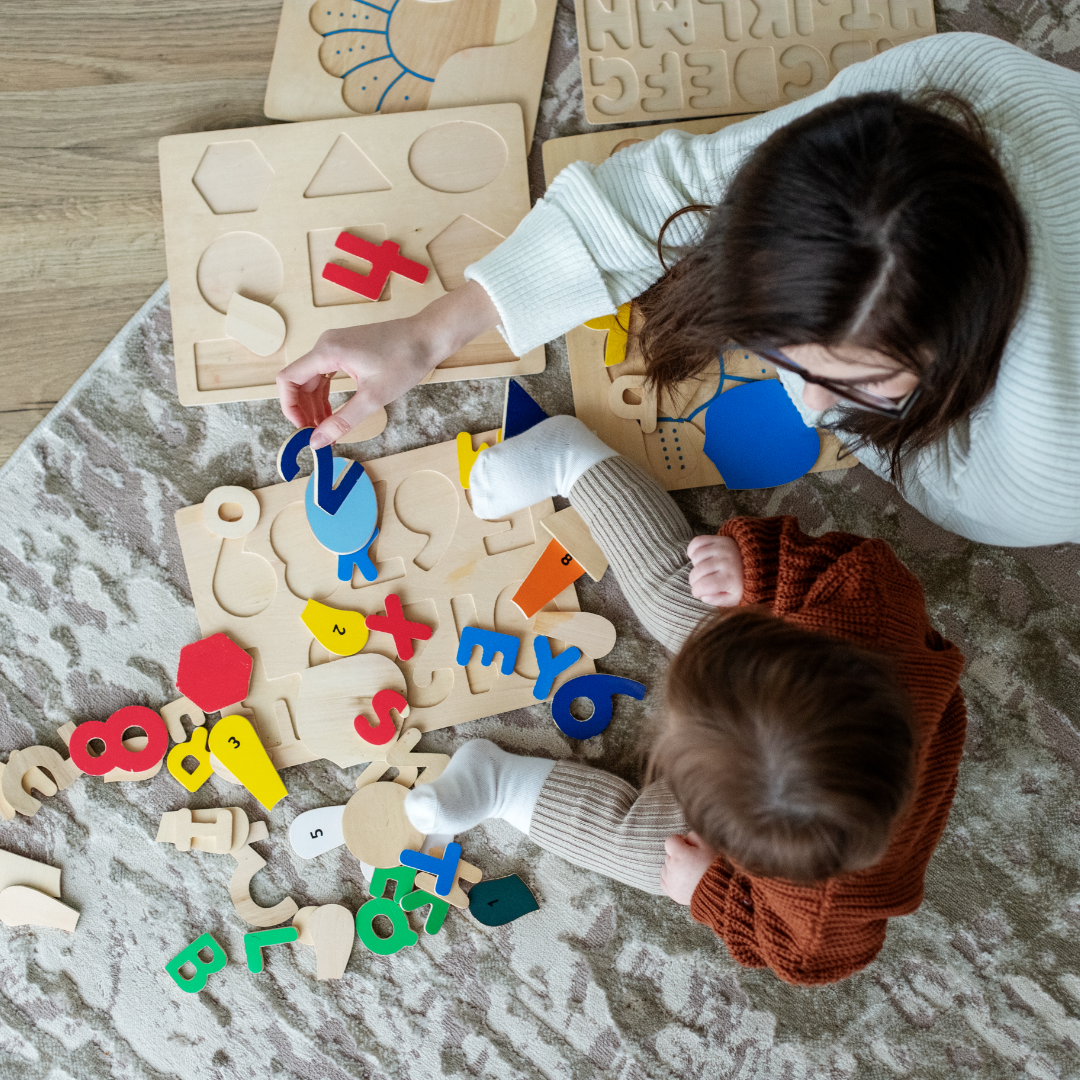 An adult and a child sit on a floor surrounded by colourful blocks in the shape of letters and numbers