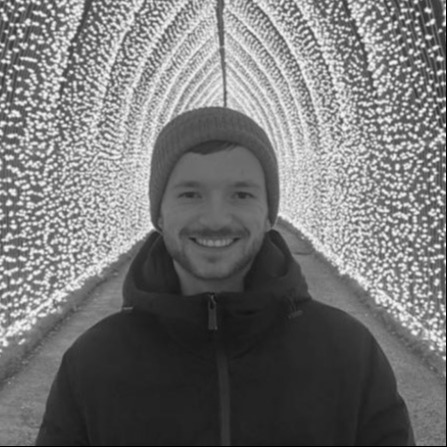 A greyscale inage of a smiling person wearing a beanie hat and a dark coloured coat. Behind them is a wall of lights rising up on either side