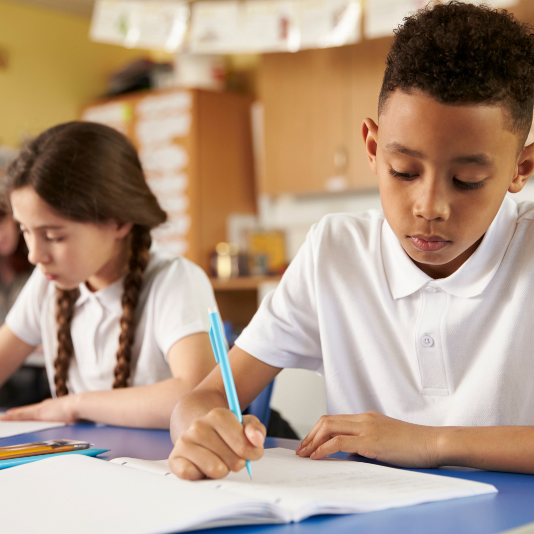 Two children sitting at a desk. They each wear a white polo shirt and the child in the foreground is writing with a blue pen.