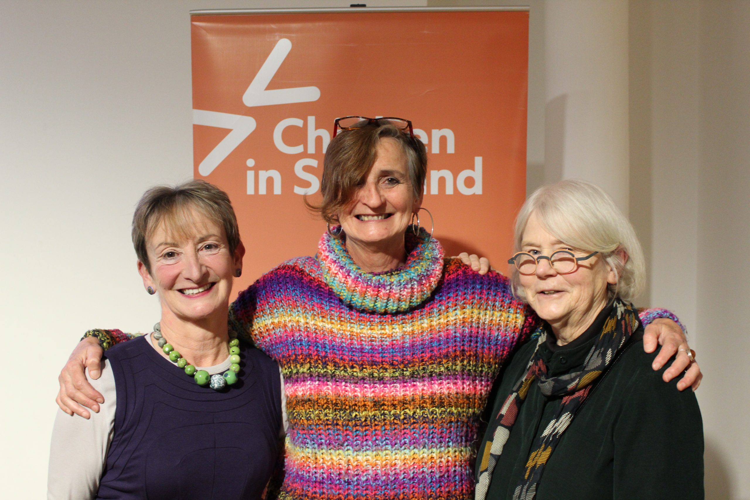 An image of three smiling people. They all have short hair. The person in the middle has their arms around the shoulders of the other two. Behind them is a banner with white text on it.