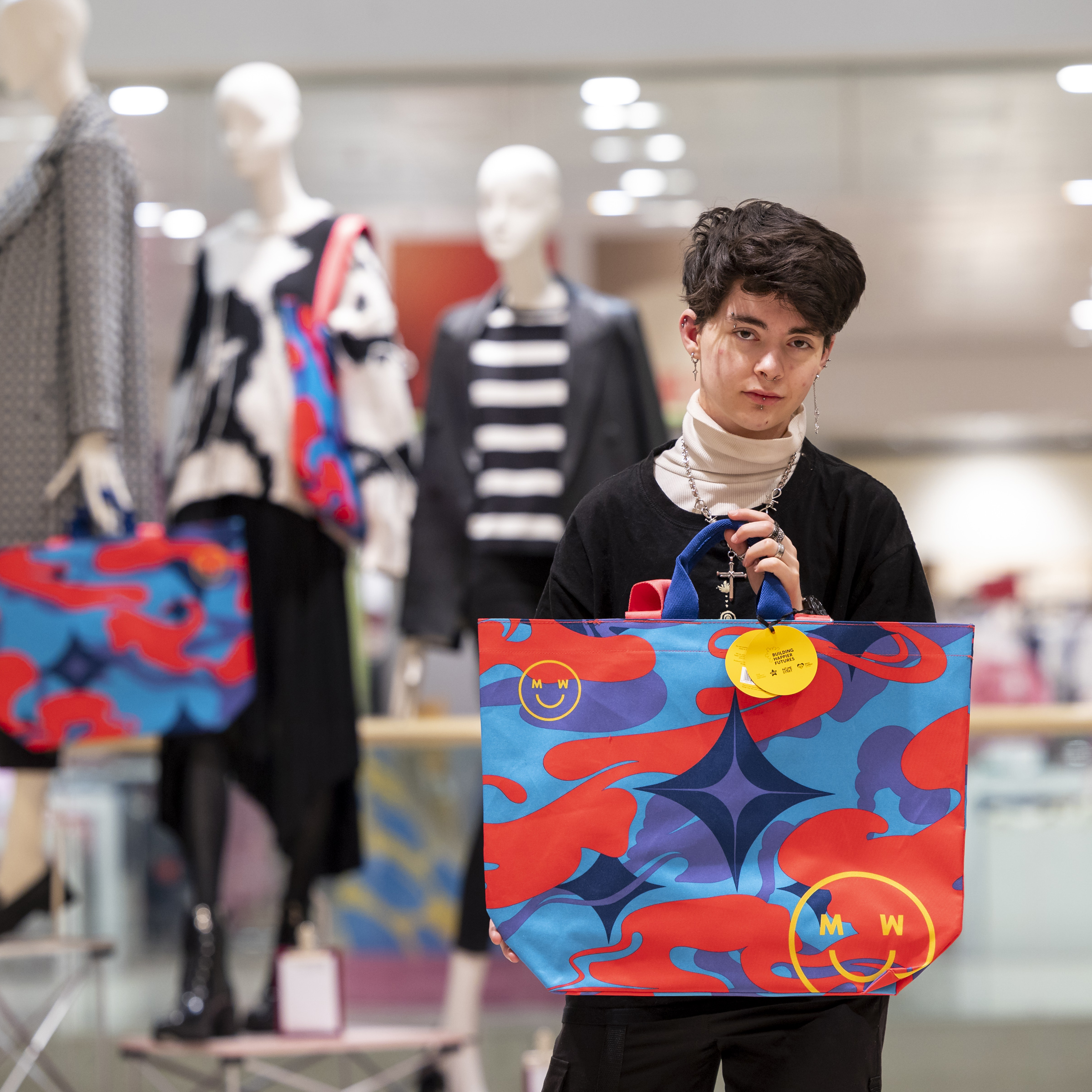 A young person with dark hair stands in front of a group of fashion mannequins. Wearing a black jumper, they hold a brightly coloured tote bag, which features swirls of blue, purple and red