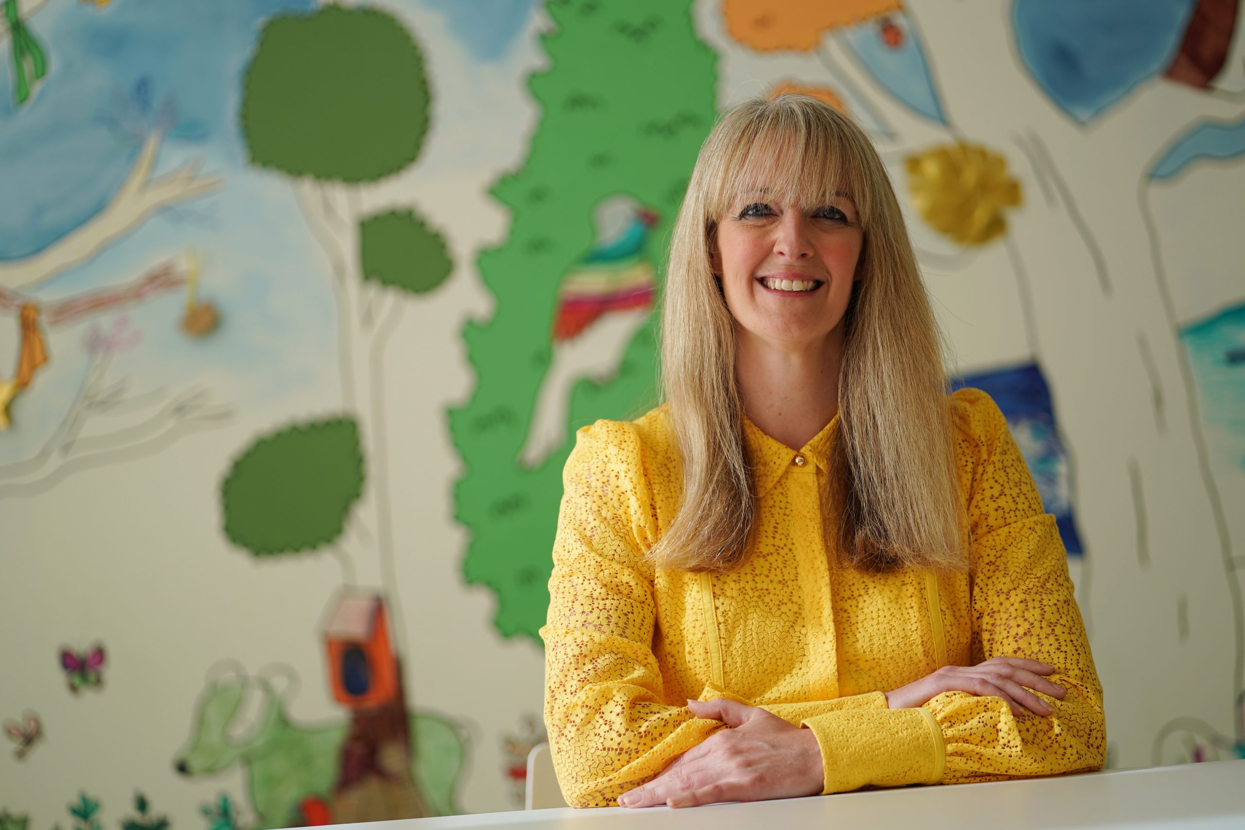 A woman with long blonde hair smiles into the camera, wearing a bright yellow shirt. Behind her is a blurred mural of trees and animals