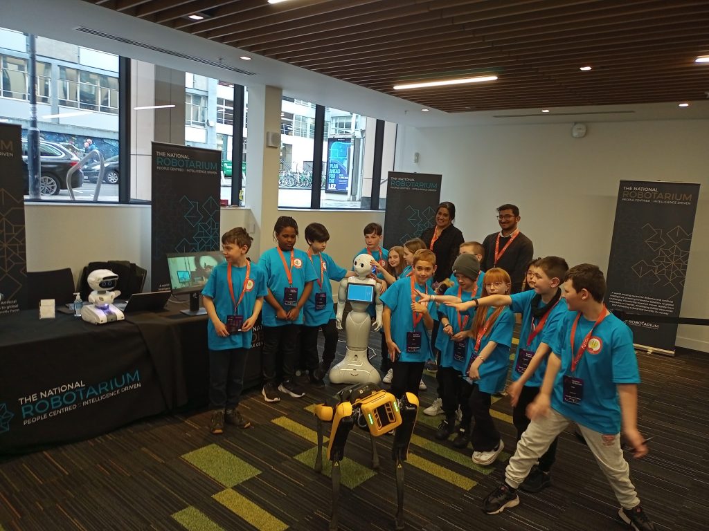 A group of children interact with an AI robot. They are all wearing blue t-shirts with orange lanyards hanging around their necks. In the background, two smiling adults supervise the children.