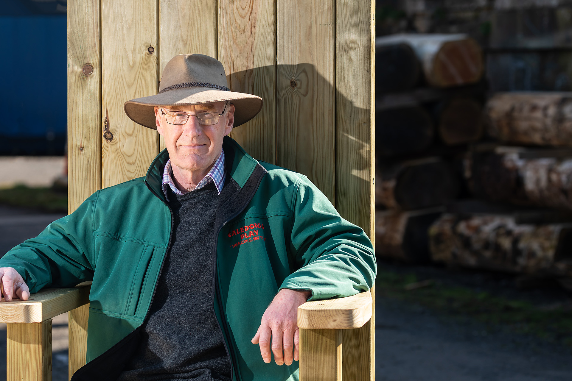 A man wearing a brown hat and green fleece jumper sits in a large outdoor wooden chair