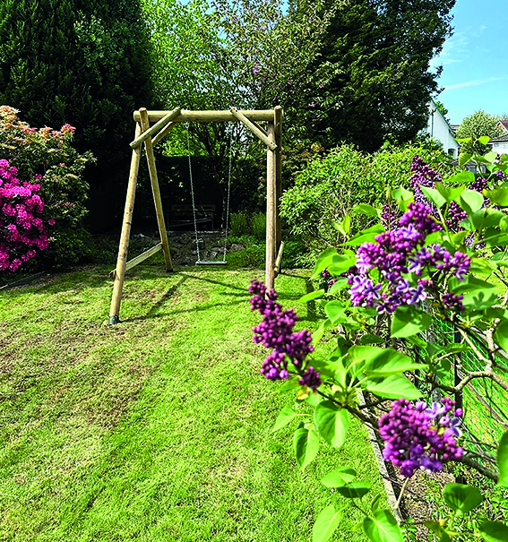 A large timber frame swing set sits in a leafy green garden bathed in sunshine. In the foreground, there are purple flowers.