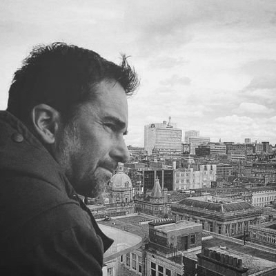 A black and white photo of the side profile of a person with short hair, standing in front of a high up view of buildings