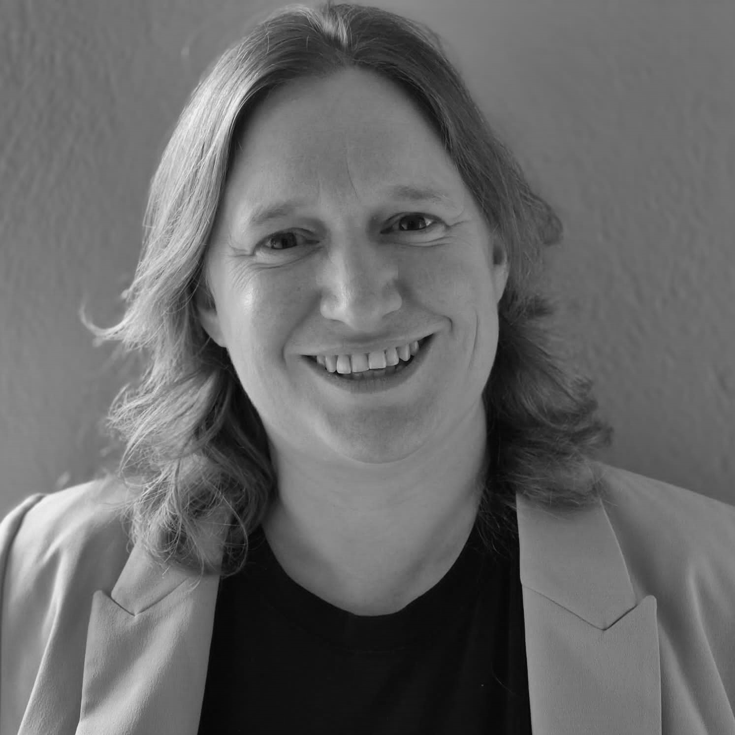 Black and white headshot of a smiling person with shoulder length hair, wearing jacket