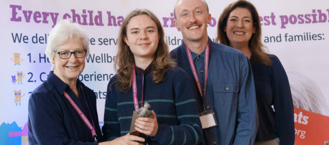 Four adults and one young person stand in a row, all smiling at the camera. In the middle, the young person holds a an award with a teddy bear on the top.