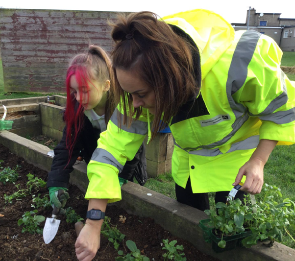 A person in a high-visibility jacet reaches into a raised planter, a child beside them holds a garden trowel. The child has pink hair.