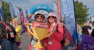 Two people in walking gear at the finish line of the Kiltwalk event, one holds cut out of a trophy