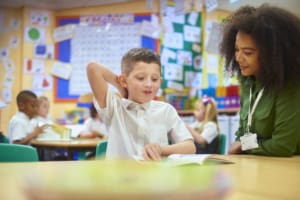 A primary aged schoolboy sits with his teacher in a one-to-one reading session. He reads aloud to her and is smiling clearly enjoying the book. The teacher is smiling too