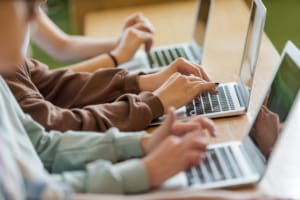 three pairs of hands rest on three laptops typing. Focus on centre person wearing brown jumper