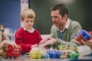 A teacher helps a young pupil in a red jumper. Egg carton crafts seen in foreground