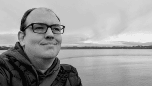 Black and white image of a person with short hair and glasses looking away from the back drop of the sea and cloudy sky